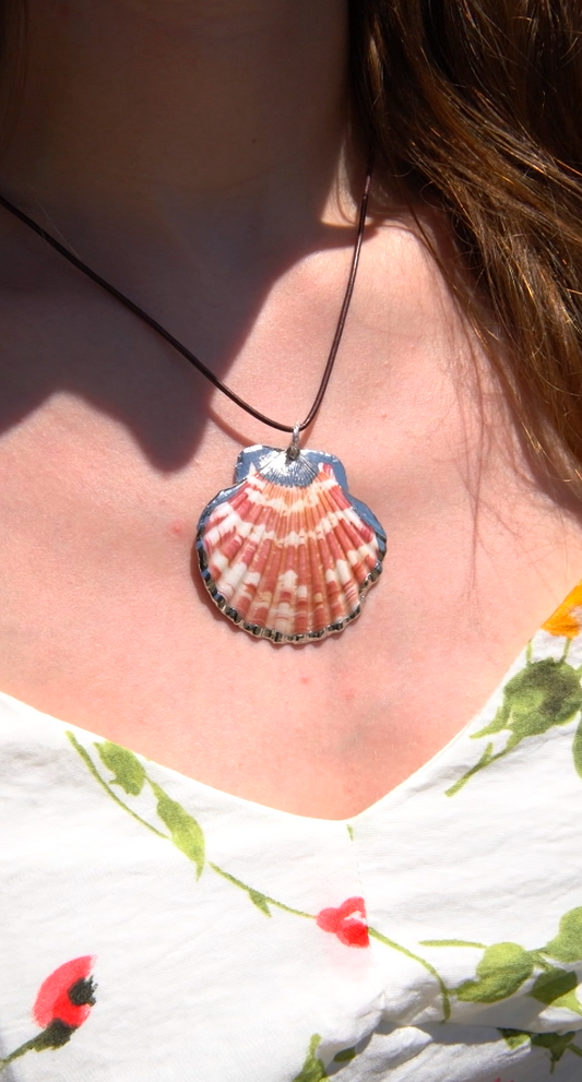 A scallop shell pendant necklace with a dark cord, worn by a person in bright sunlight. The pendant features natural pink, orange, and white striped patterns with a blue-gray edge.